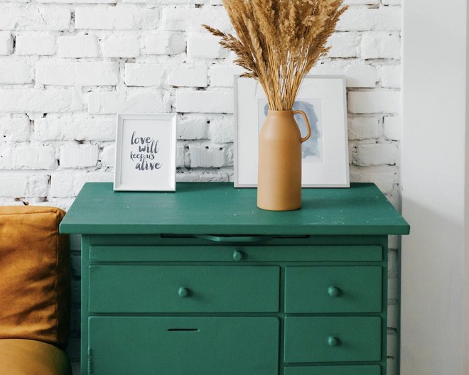 A green chest of drawers with framed photos and a brown vase on top, against a white brick wall.